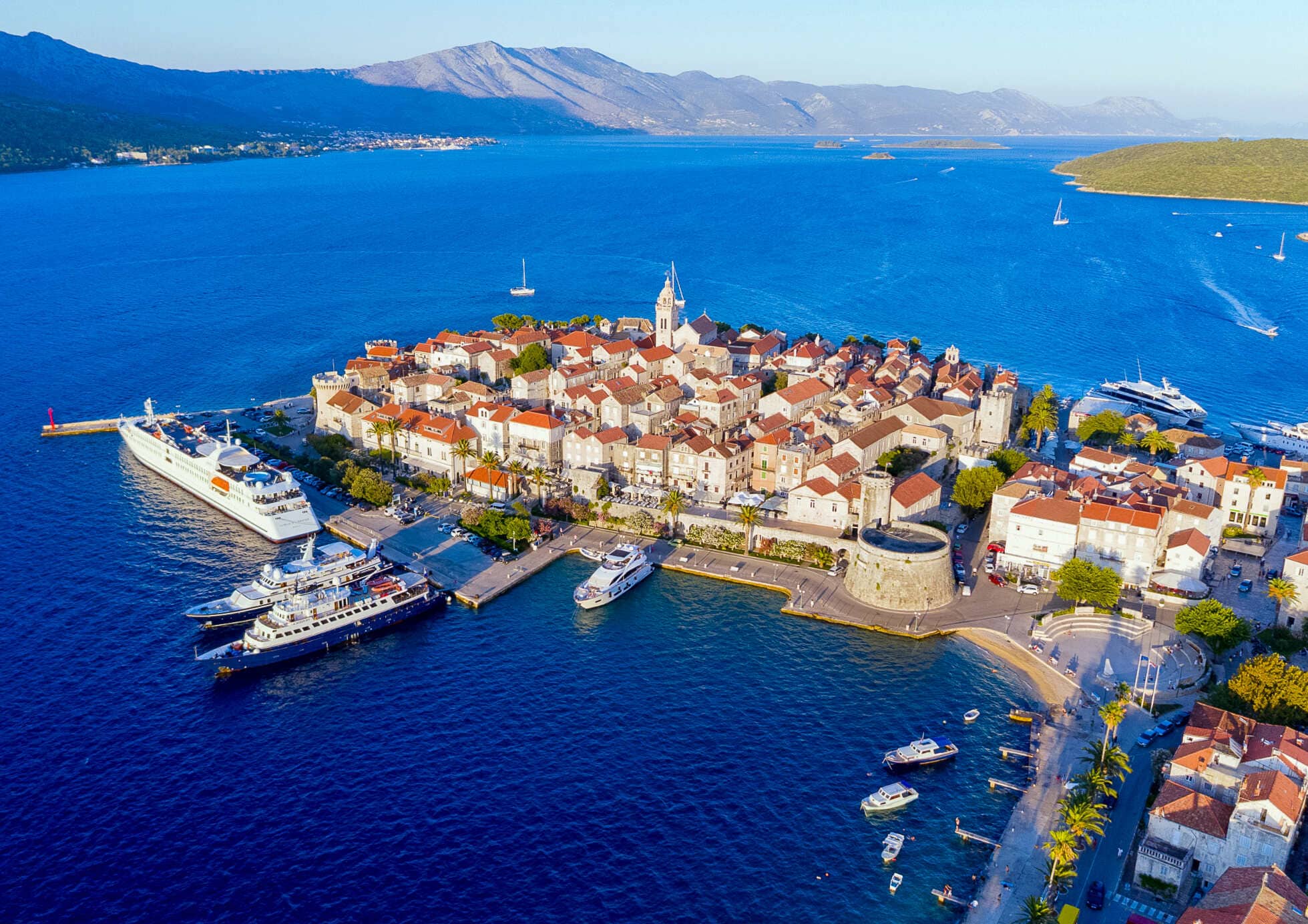 Aerial view of Korčula Old Town in Croatia, showing its historic stone buildings, red-tiled roofs, waterfront harbor, boats, and surrounding deep-blue Adriatic Sea with mountains in the distance.