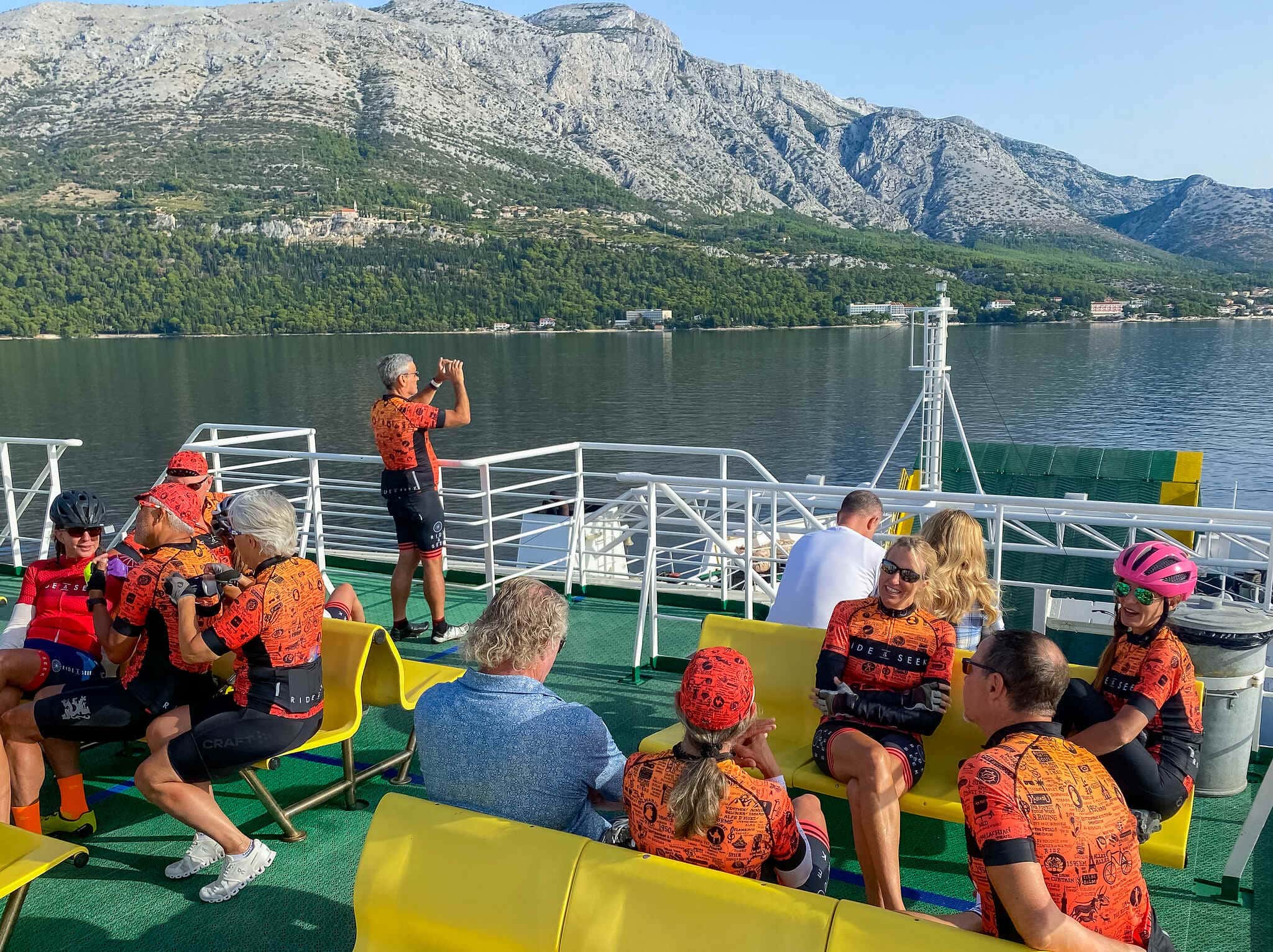 Ride & Seek cyclists relaxing on a ferry deck, with Montenegro’s dramatic limestone mountains and forested shoreline rising above the calm waters of the Bay of Kotor.