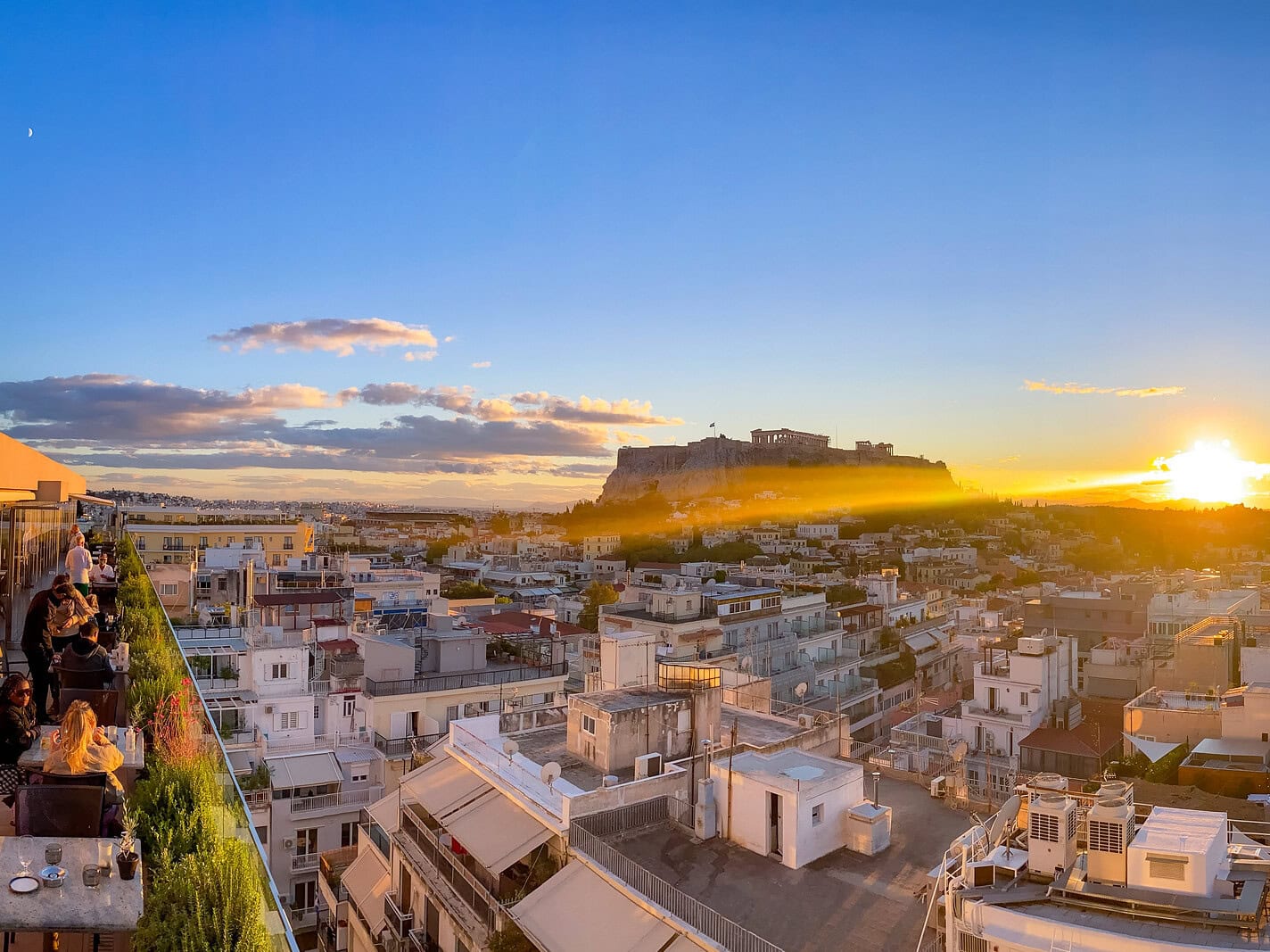 Rooftop terrace overlooking Athens at sunset, with the illuminated Acropolis and Parthenon rising above the city skyline.