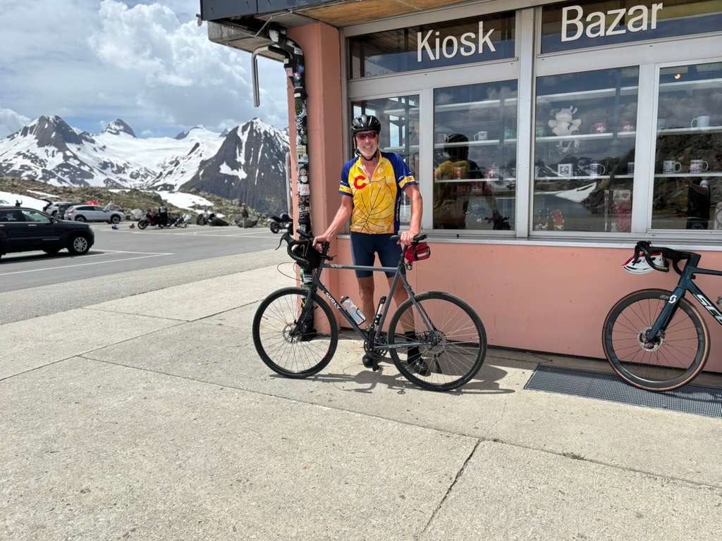 Paul standing with his bike outside a souvenir kiosk at the Nufenen Pass summit on a Ride & Seek cycling tour