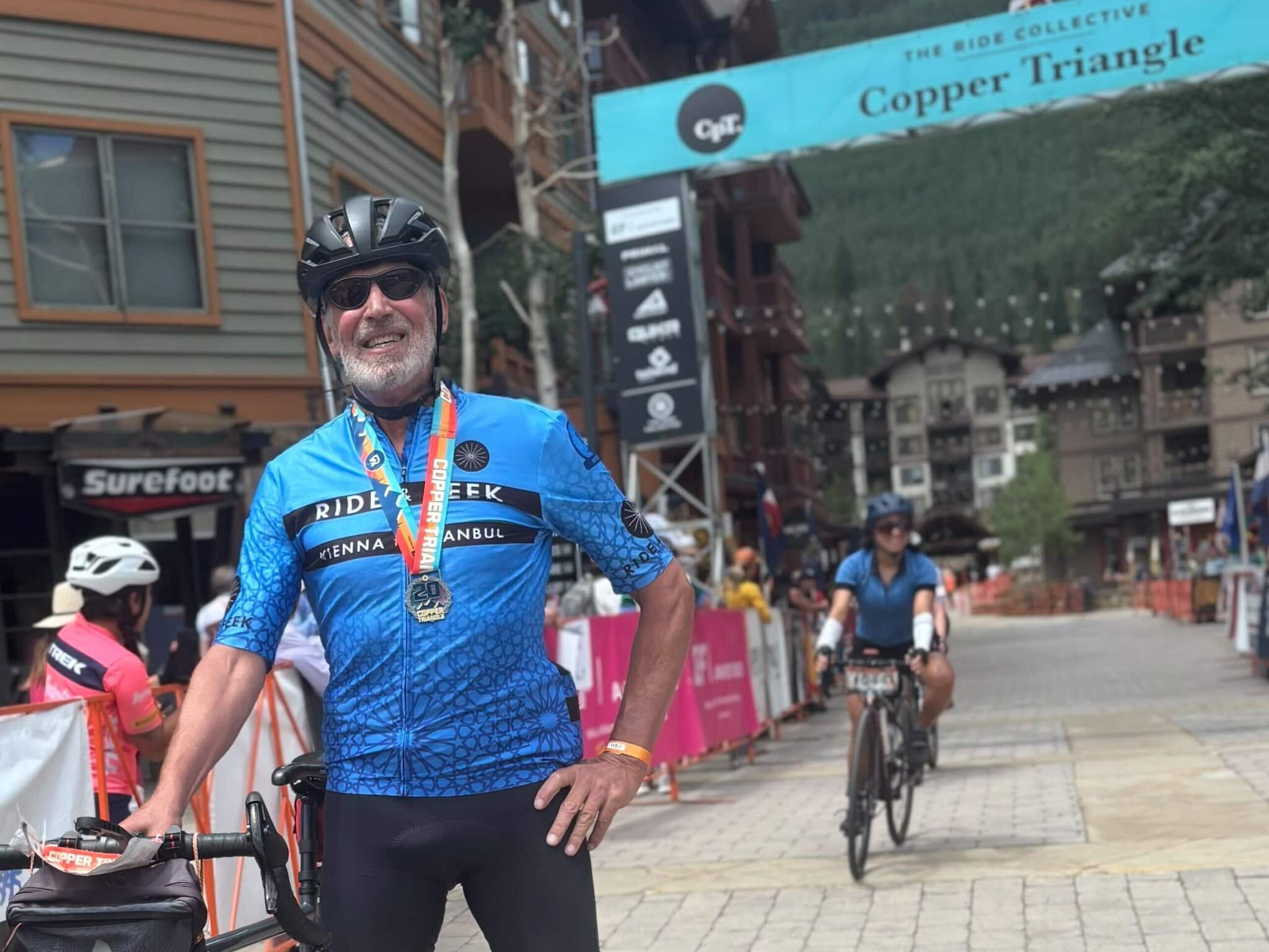 Paul standing with his bike under a Copper Triangle finish banner wearing a blue Ride and Seek cycling tours jersey and medal