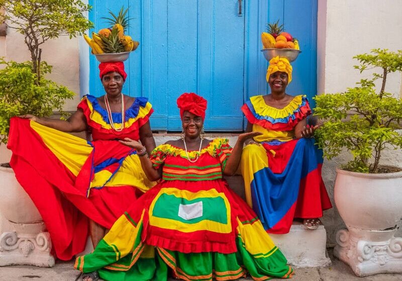 Three women in vibrant traditional dresses, two with bowls of fruit on their head dresses, smiling in front of a bright blue doorway.