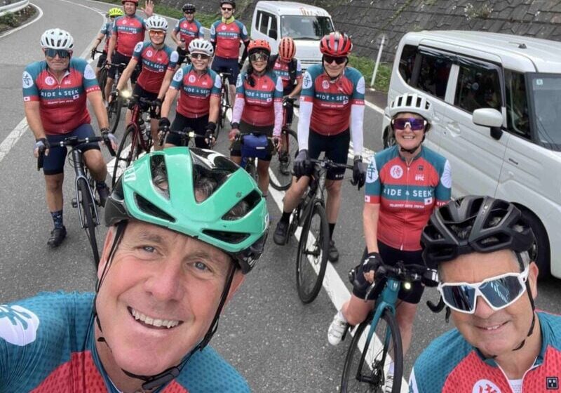 A group of cyclists wearing matching red, teal, and white "Ride & Seek" jerseys and helmets, taking a group selfie on a paved road lined with a white van and concrete retaining wall.