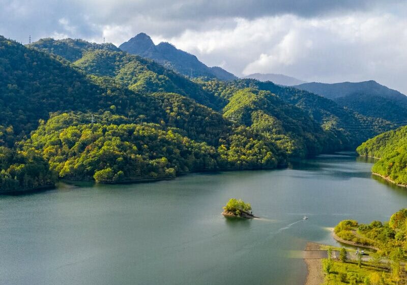 Tranquil lake bordered by forested hills and mountains, with soft sunlight highlighting the green forest and grey clouds drifting across the sky.