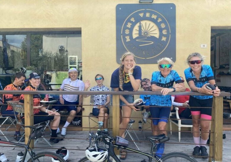 A group of three people dressed in cycling gear stand leaning on a balcony rail. another group sit on seating behind them.