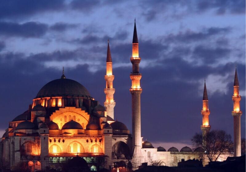 A mosque is lit up, with an evening sky behind.