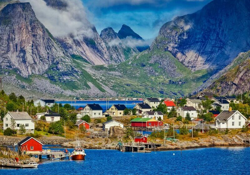 Norsemen Norway Cover Photo A picturesque Norwegian fishing village with colourful houses and boats by the shore, surrounded by dramatic mountains and low-hanging clouds.