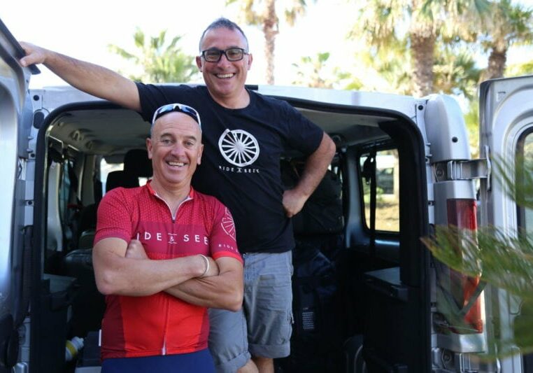 Two smiling guides wearing Ride and Seek uniforms pose naturally in front of the back of an open tour support van.